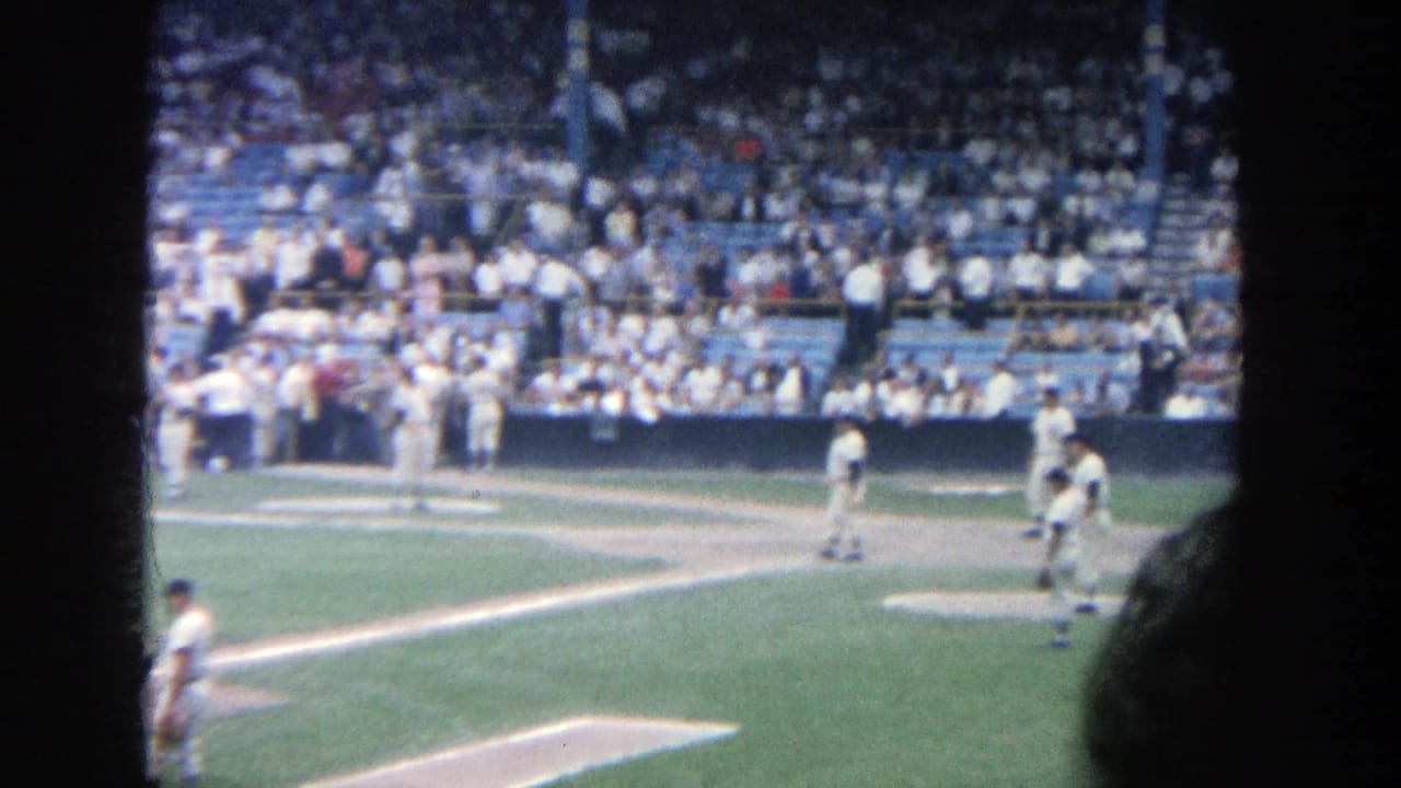 1964 Yankee Stadium home-movie frame showing players in white uniforms paused near the infield with a full crowd behind them.