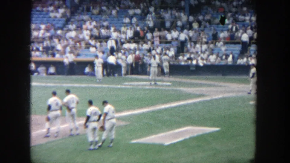 1964 Yankee Stadium frame with players in white uniforms gathered near the baseline during a pause in play.