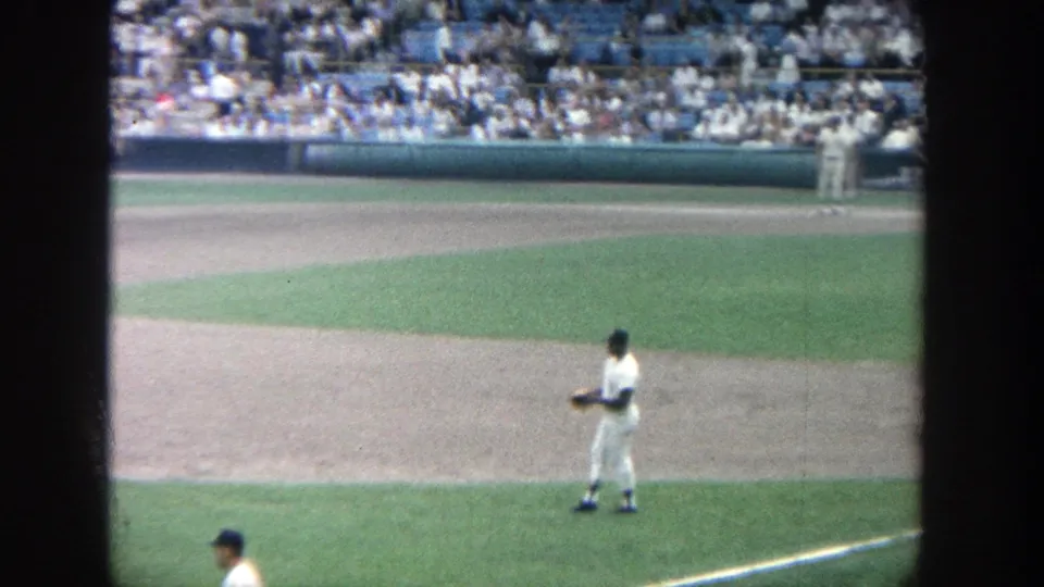 1964 Yankee Stadium frame with a baseball player near the infield edge, green turf, dirt baseline, and seated crowd beyond.