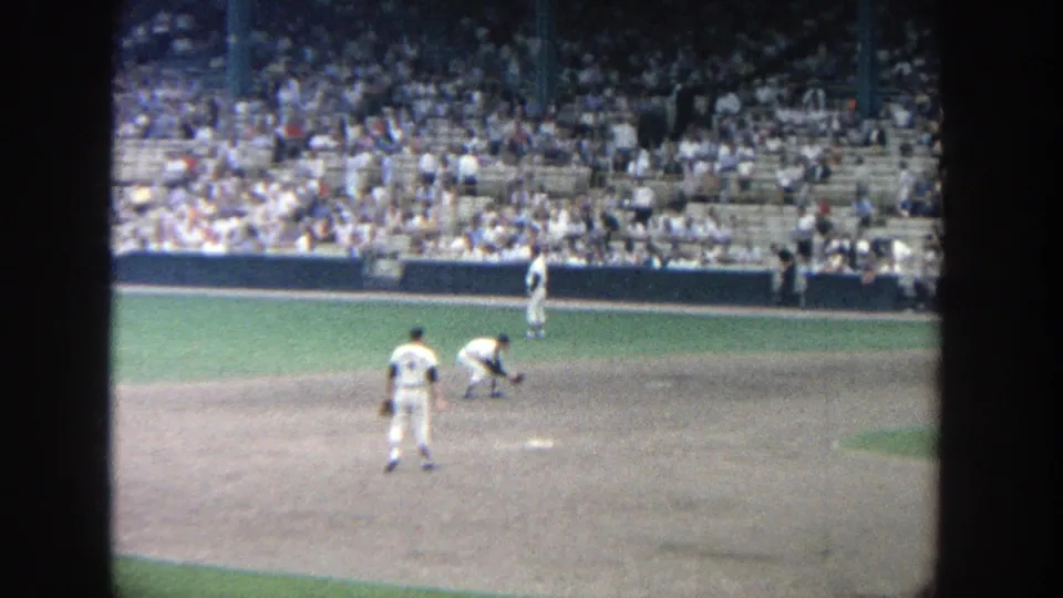 1964 Yankee Stadium home-movie frame showing baseball players on the dirt infield before a packed stand.