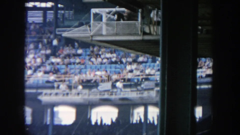 1964 Yankee Stadium home-movie frame showing spectators, railings, and upper-deck structure during the baseball game.