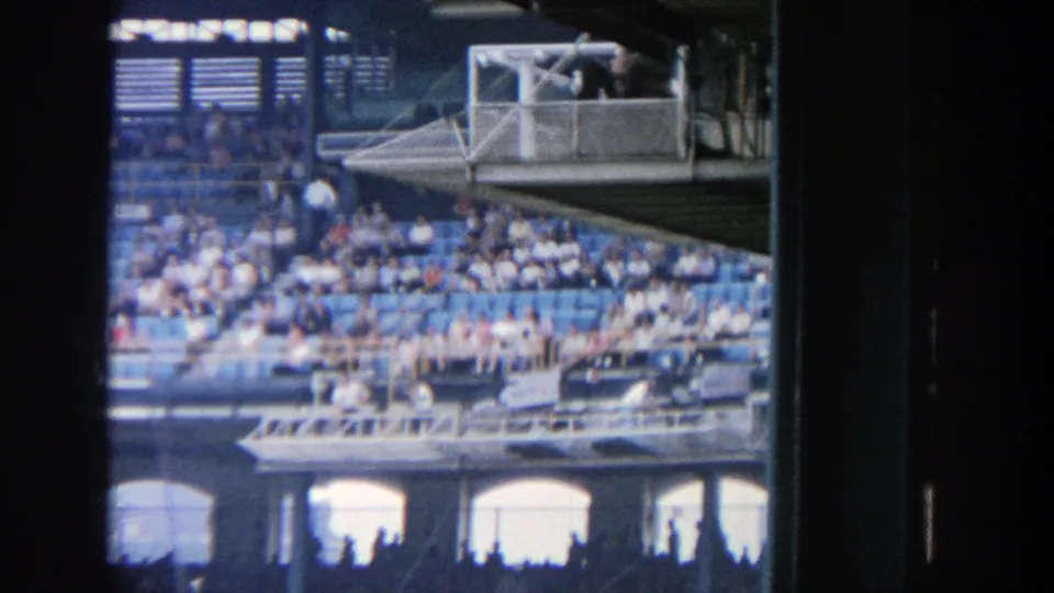 Home-movie view across Yankee Stadium seating in 1964, with dense spectators, blue seats, railings, and shaded foreground supports.