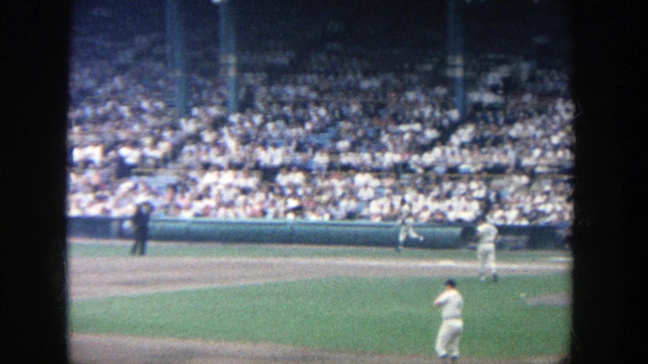 1964 Yankee Stadium view from behind home plate, with white-uniformed players on the field and a packed crowd in green bleachers.