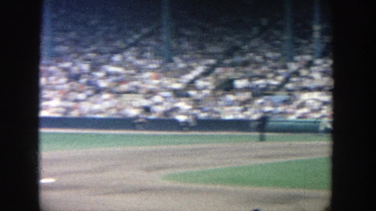 Wide 1964 Yankee Stadium ballpark view showing infield dirt, green outfield grass, distant figures and crowded stadium seating.