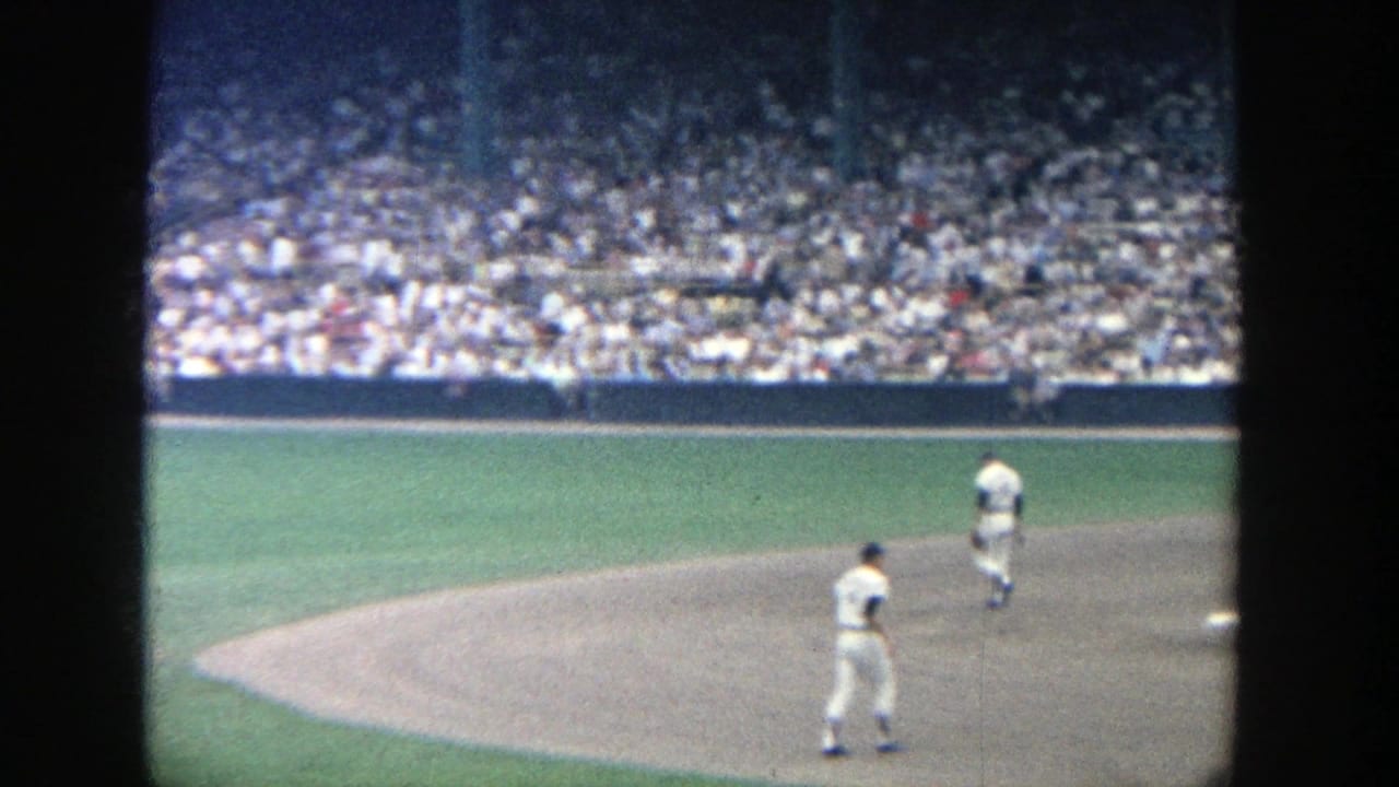 Two white-uniformed players move across the Yankee Stadium infield as spectators fill the bleachers in the background.