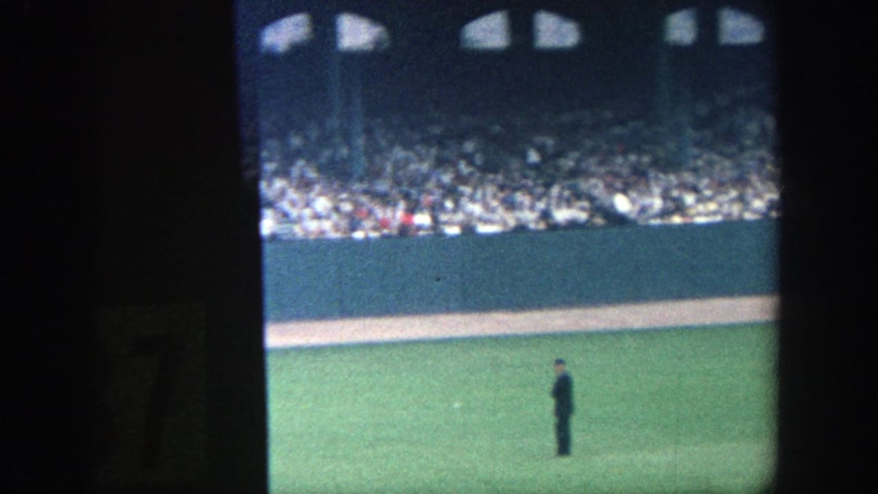 Wide outfield view with a dark-clad figure on the grass and spectators behind the wall.
