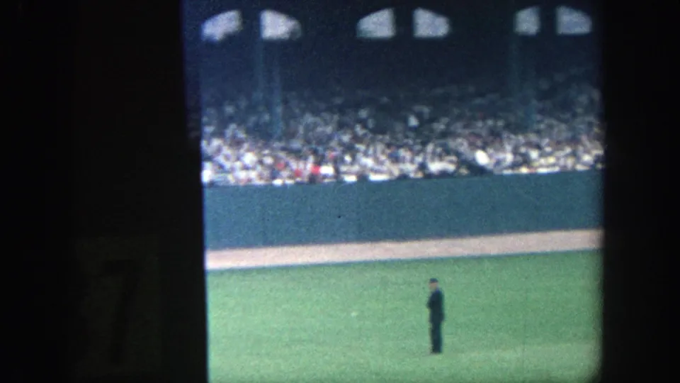 1964 Yankee Stadium frame with a dark-uniformed field figure in the outfield and a dense spectator crowd behind the wall.