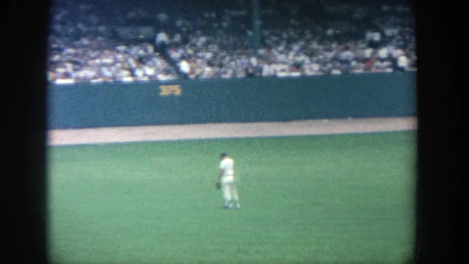 1964 Yankee Stadium 8mm frame showing a baseball player on the outfield grass below a crowded stand and outfield wall.
