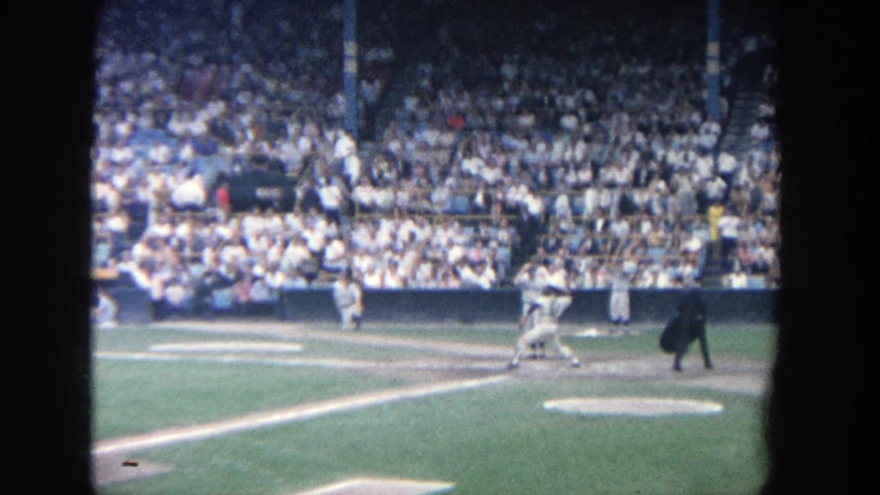 1964 Yankee Stadium home-movie frame showing players near home plate as spectators fill the stands behind the field.