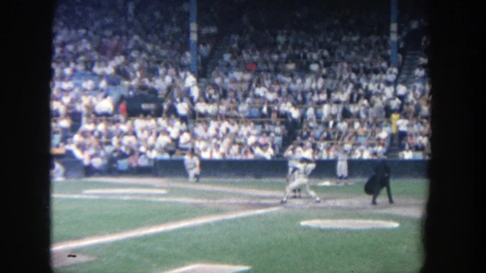 1964 Yankee Stadium home-movie frame showing players near home plate as spectators fill the stands behind the field.