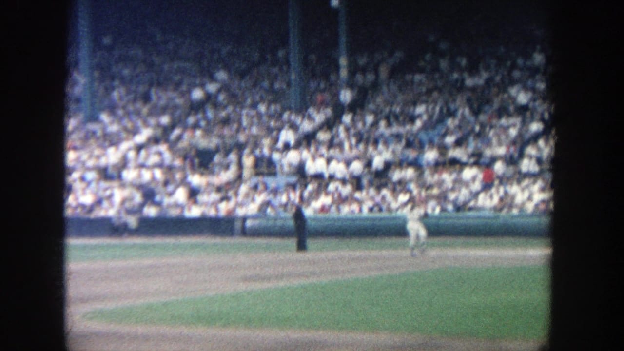 1964 Yankee Stadium home-movie frame showing a distant field view, players in white uniforms, and a large seated crowd.