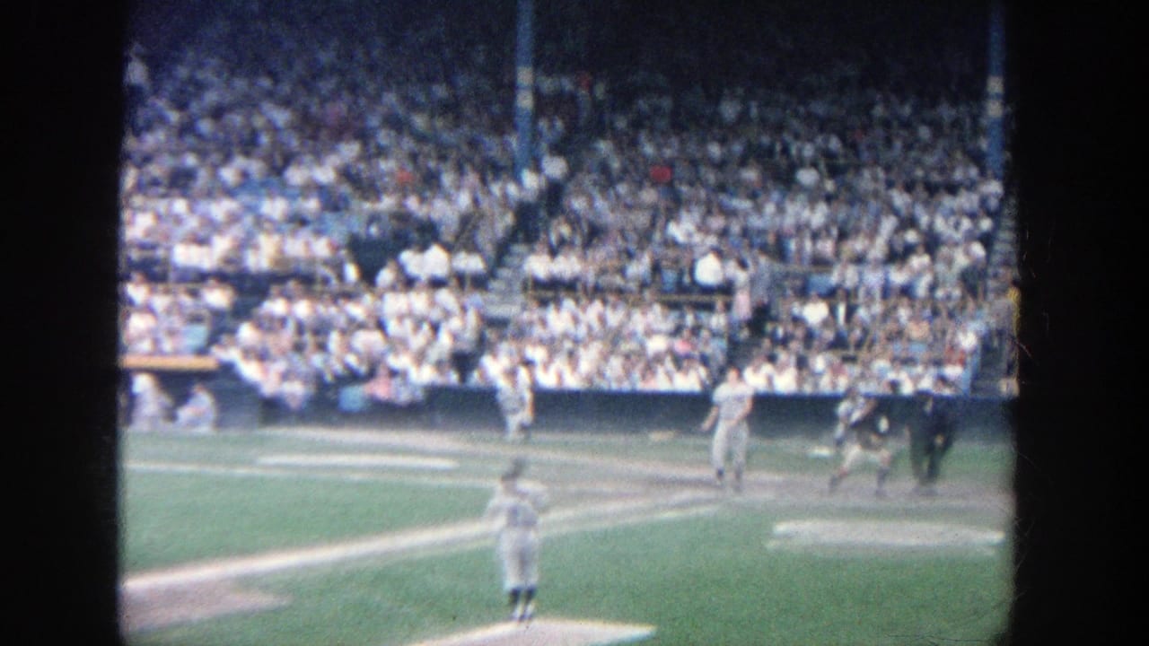 1964 Yankee Stadium spectator-shot frame showing home plate action with players and umpires before crowded seating.