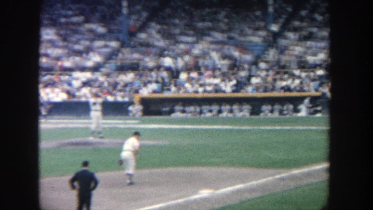 1964 Yankee Stadium frame showing the pitcher on the mound, an official in the foreground, and spectators beyond the dugout.