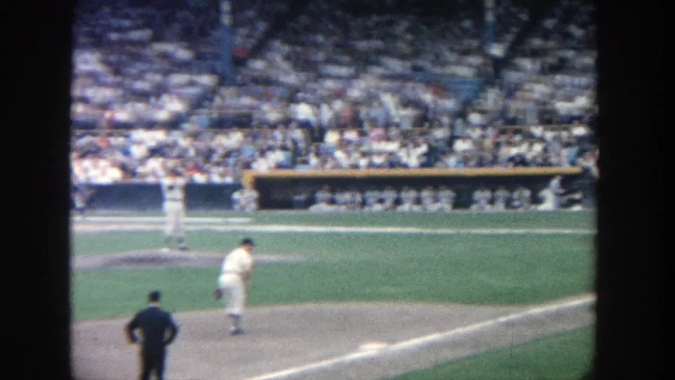 1964 Yankee Stadium frame showing the pitcher on the mound, an official in the foreground, and spectators beyond the dugout.