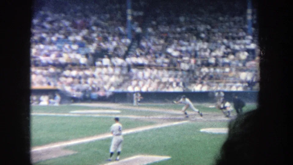 1964 Yankee Stadium home-movie frame showing a batter swinging, with pitcher, catcher, umpire, and stands behind.