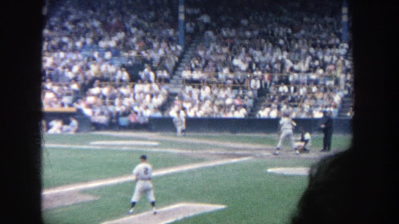 Batter, catcher, umpire, and fielders visible with crowded stadium seats behind them.