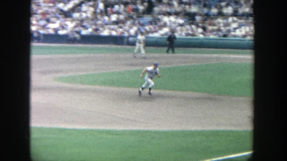 1964 Yankee Stadium home-movie frame showing a baseball player running on the infield as spectators fill the background stands.
