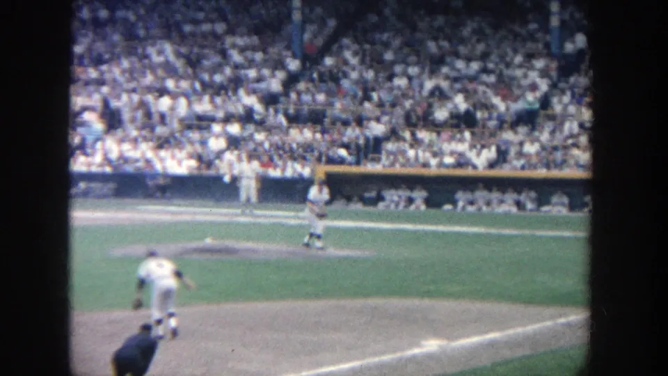 1964 Yankee Stadium home-movie frame showing the pitcher on the mound, another player in the foreground, and spectators beyond the dugout.