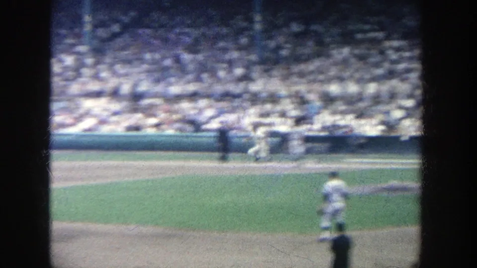 1964 Yankee Stadium home-movie frame with motion blur, a player near the lower field, infield dirt, and a dense spectator crowd behind.