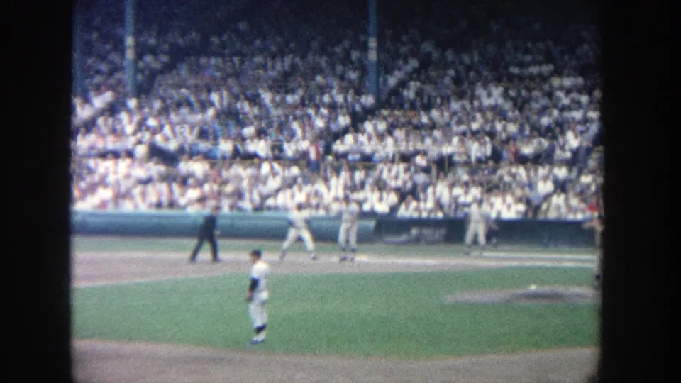 Spectator-shot 1964 Yankee Stadium frame with players, an umpire and a packed crowd behind the infield.