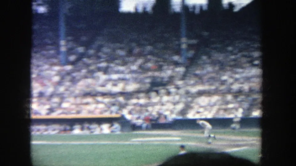 1964 Yankee Stadium home-movie frame showing a player running near first base with spectators in the background.