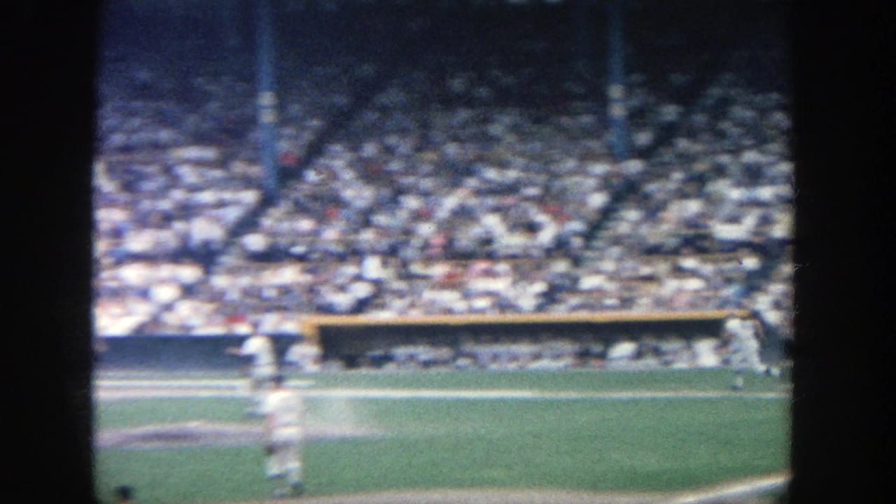 1964 8mm view of a baseball game at Yankee Stadium, with players near the infield and crowded stands in the background.