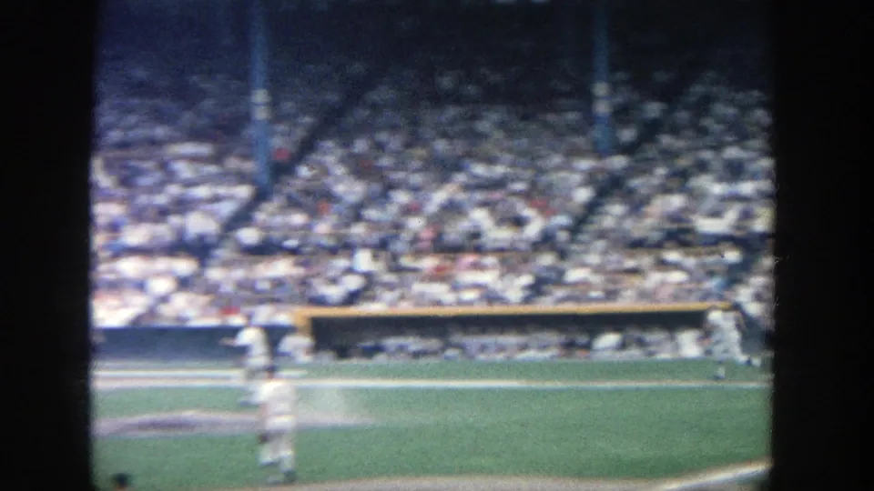 1964 8mm view of a baseball game at Yankee Stadium, with players near the infield and crowded stands in the background.