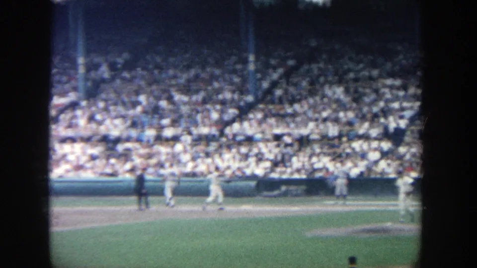 Wide 8mm frame of the Yankee Stadium baseball diamond, with players near the baseline and a packed audience in the seats.