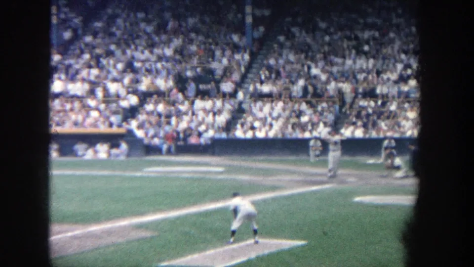 1964 Yankee Stadium home-movie frame showing a batter or runner moving up the first-base line as players and spectators remain visible.