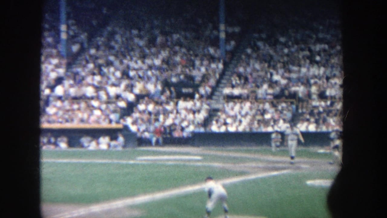 1964 Yankee Stadium home-movie frame showing a packed grandstand, infield lines, and baseball players seen from the stands.