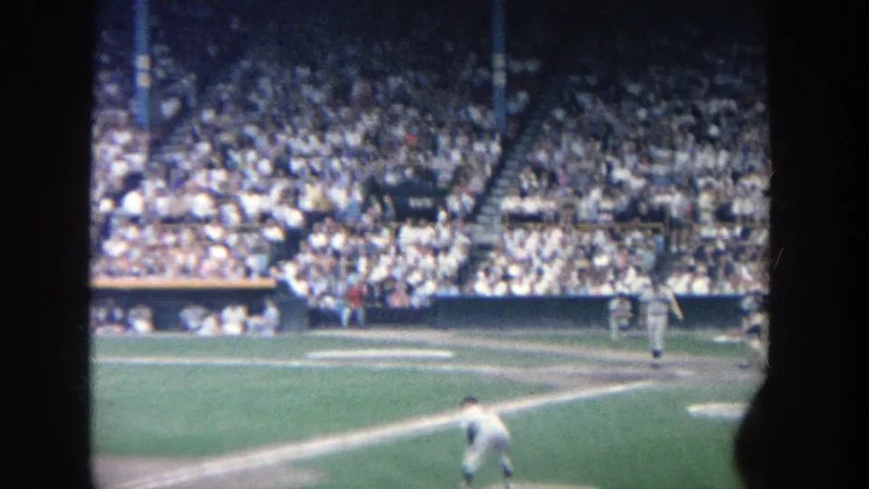 1964 Yankee Stadium home-movie frame showing a packed grandstand, infield lines, and baseball players seen from the stands.