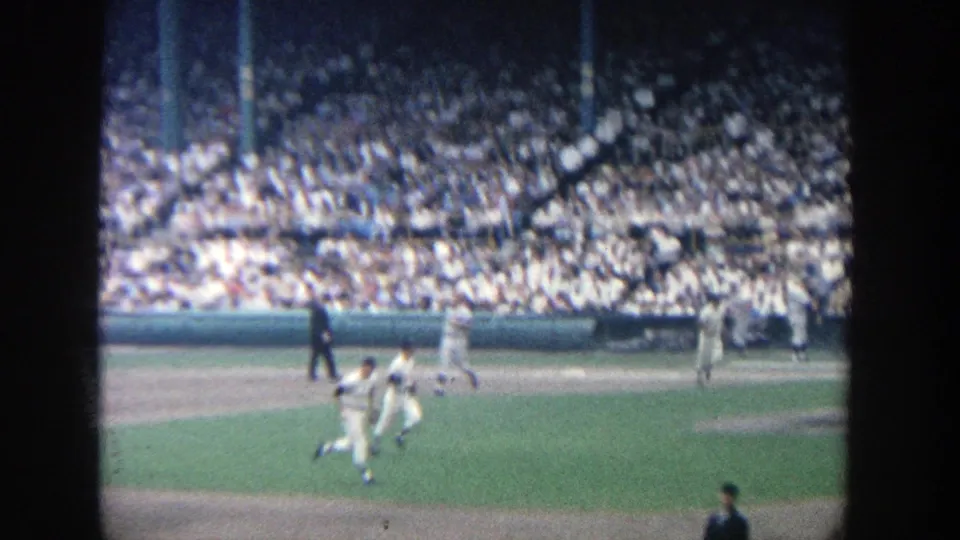 1964 Yankee Stadium home-movie frame showing baseball players moving on the infield in front of a large crowd.