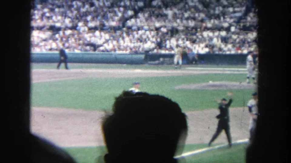 1964 8mm stadium view with an official signaling near the line, players on the field, and a spectator silhouette at the bottom edge.