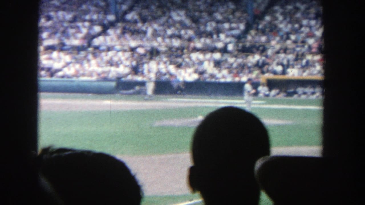 Baseball field seen from the stands with crowd, players, and dark foreground silhouettes.
