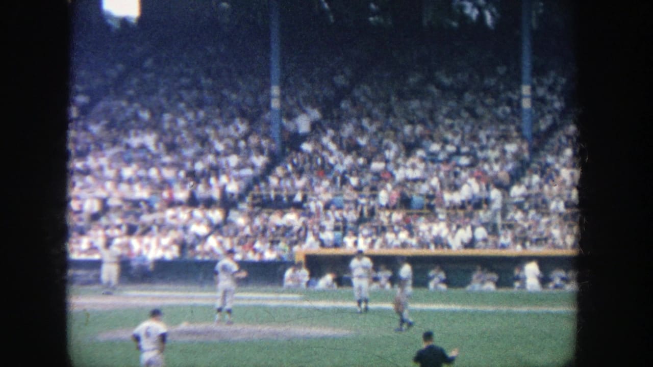 Players stand near the infield as a packed stadium crowd fills the background.