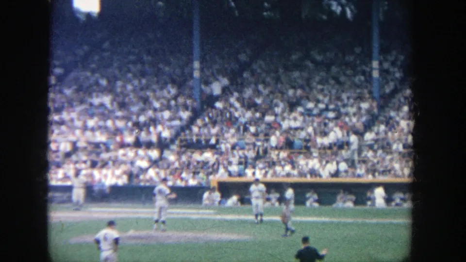 Wide 8mm shot of players on the Yankee Stadium field with crowded stands and dugout areas visible behind them.