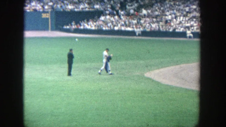 1964 Yankee Stadium home-movie frame showing a player crossing the grass near an official, with outfield wall and spectators beyond.