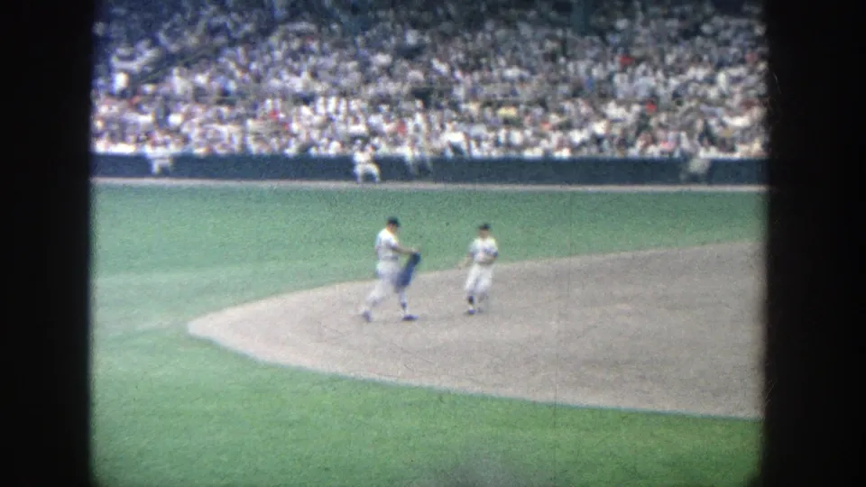 1964 Yankee Stadium home-movie frame showing two uniformed players on the infield as one appears to handle a jacket.
