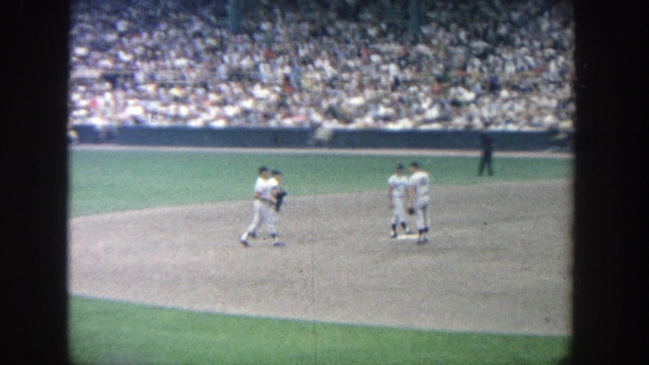 1964 Yankee Stadium home-movie frame showing players in white uniforms on the infield before play, with packed stands behind them.