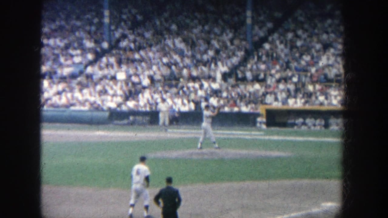 A pitcher stands near the mound while players and an umpire-like figure are visible before the crowd.