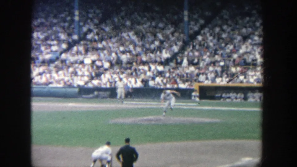 Home-movie view at Yankee Stadium in 1964 showing a pitcher winding up, with a player and official-like figure in the foreground.