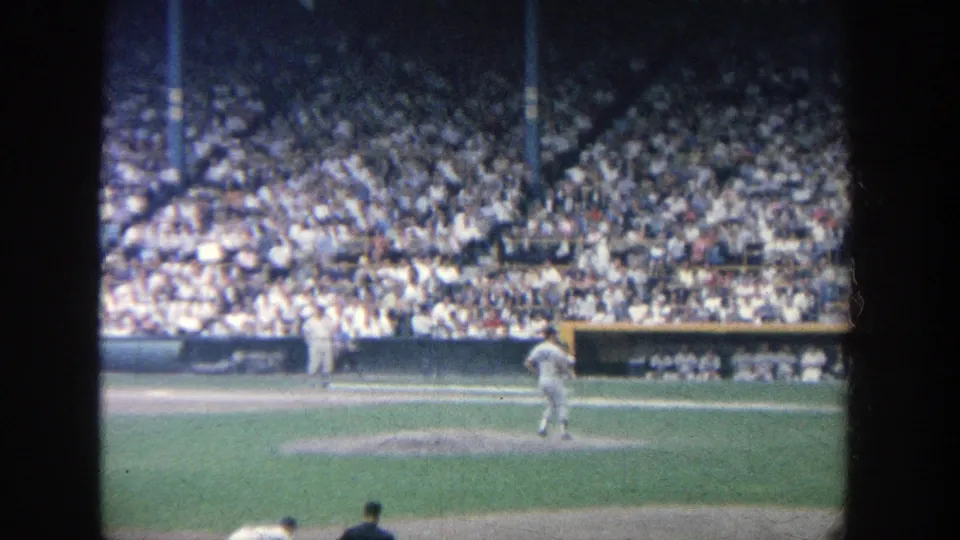 1964 Yankee Stadium frame showing a pitcher after release on the mound, with crowded seating and a dugout area in the background.