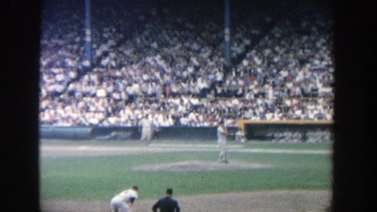 Baseball game seen from the stands with pitcher, fielders, and a crowded grandstand.