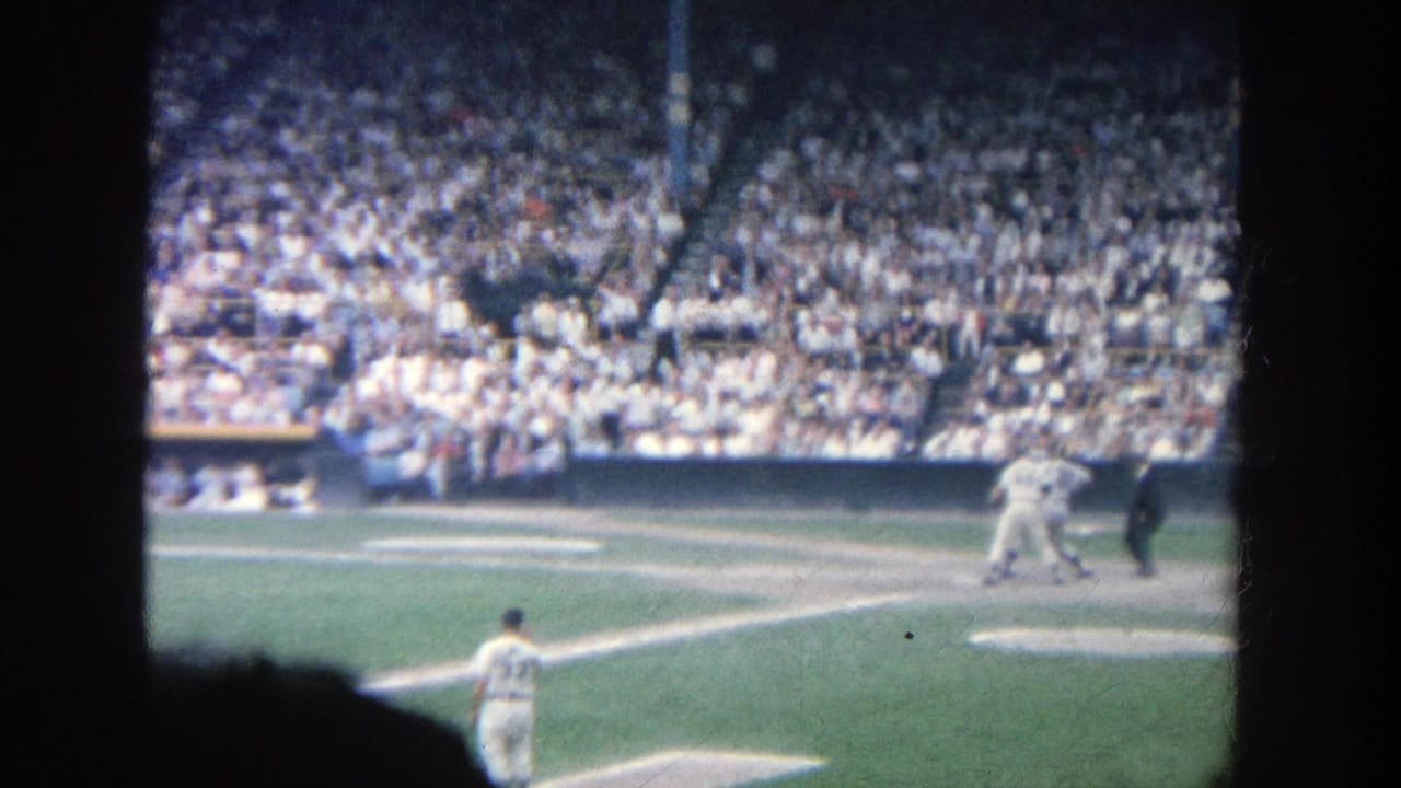 Batter at home plate with pitcher in foreground and crowded stadium seats behind.