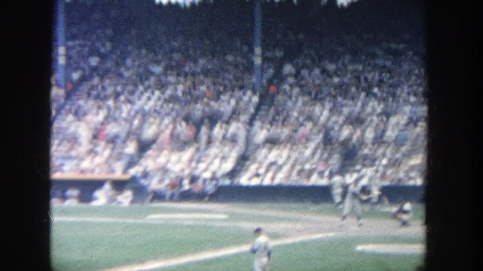 Soft 8mm frame from a 1964 Yankee Stadium game, showing the at-bat area, infield grass, and packed spectator seating.