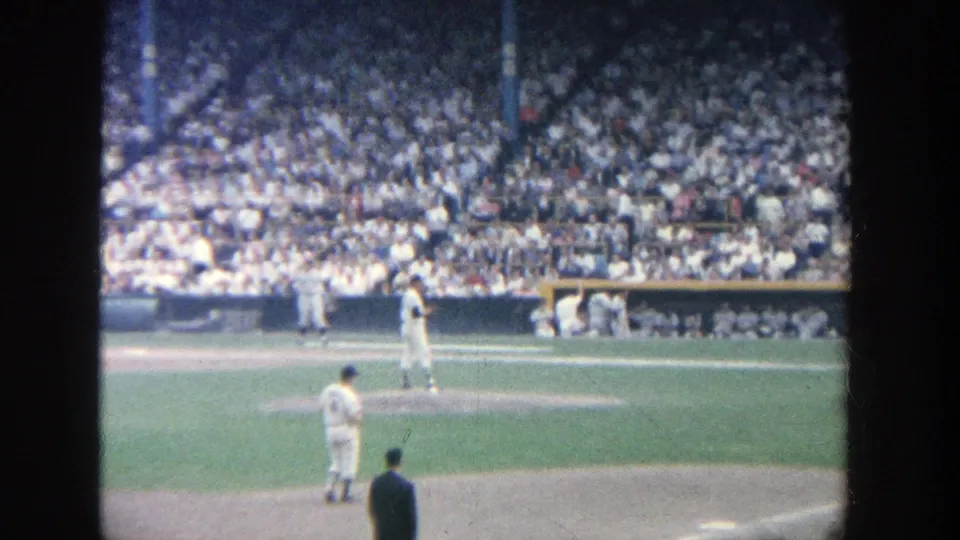 1964 Yankee Stadium home-movie frame showing a pitcher set on the mound, a batter near home plate, and packed stands in the background.