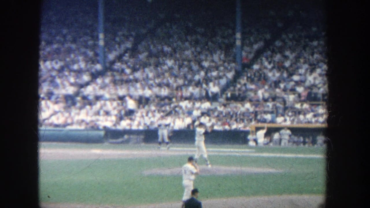 Pitcher faces home plate as a batter waits, with the dugout and full stands beyond the field.