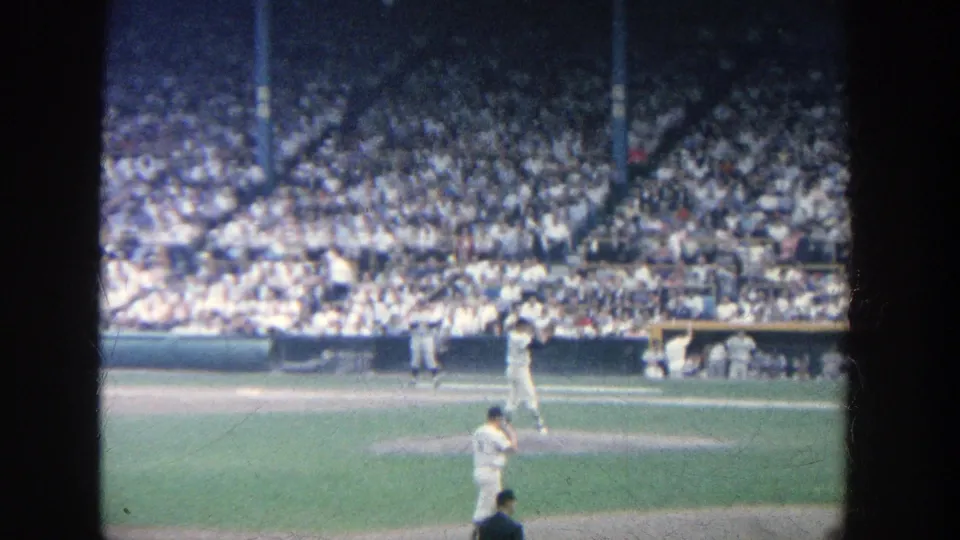 Spectator-shot 1964 Yankee Stadium frame with pitcher, batter, field-level dugout area, and dense grandstand crowd visible.