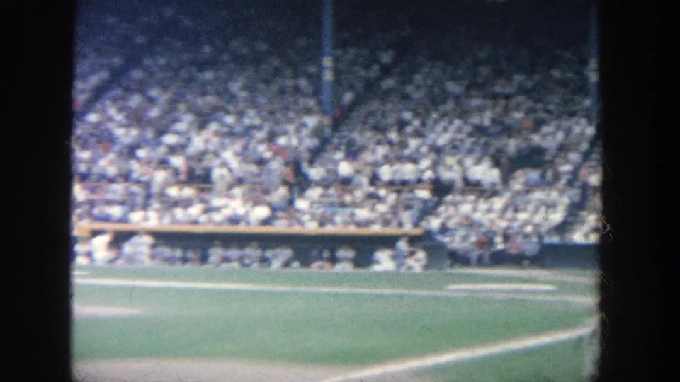 1964 Yankee Stadium home-movie frame showing field grass, infield edge, dugout area, and a packed grandstand from a spectator angle.