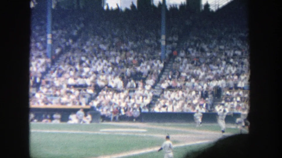 Late-frame 1964 Yankee Stadium view with a player near the lower field edge, crowded stands, dugout area, and visible film-frame border.